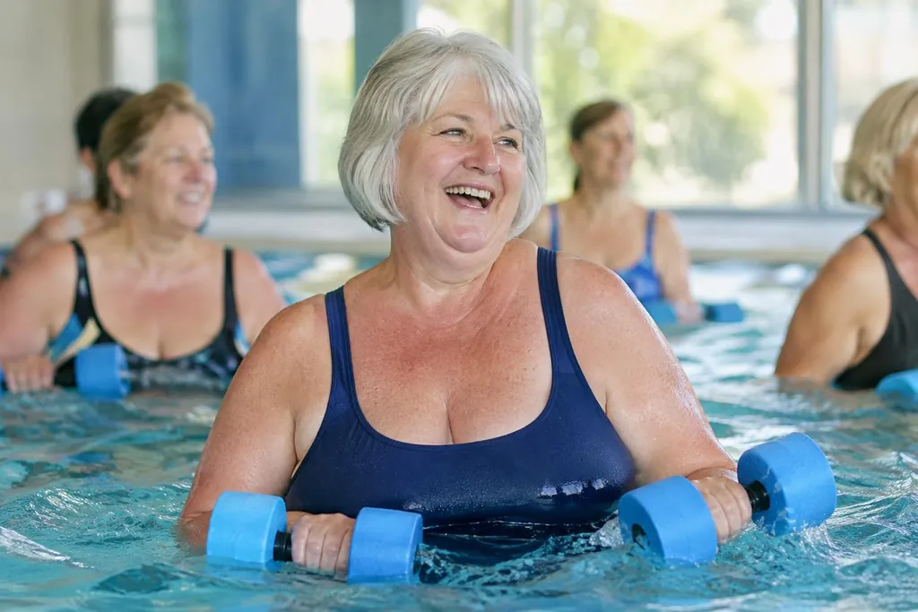 Femme d'environ soixante-cinq ans en maillot une pièce bleu marine qui rit pendant un cours d'aquagym, haltères en mousse en main