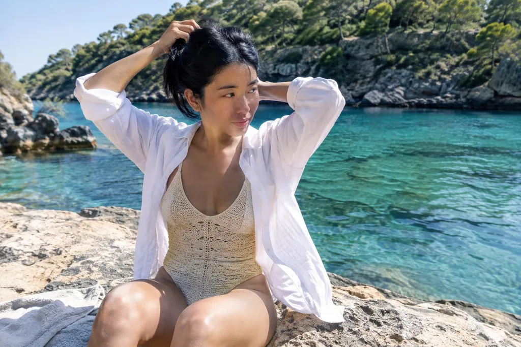Jeune femme en bikini crochet écru doublé, assise sur un rocher plat dans une crique, qui attache ses cheveux