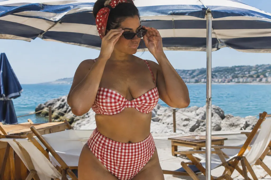 Femme d'une trentaine d'années en bikini vichy rouge à culotte haute qui ajuste ses lunettes sous un parasol rayé