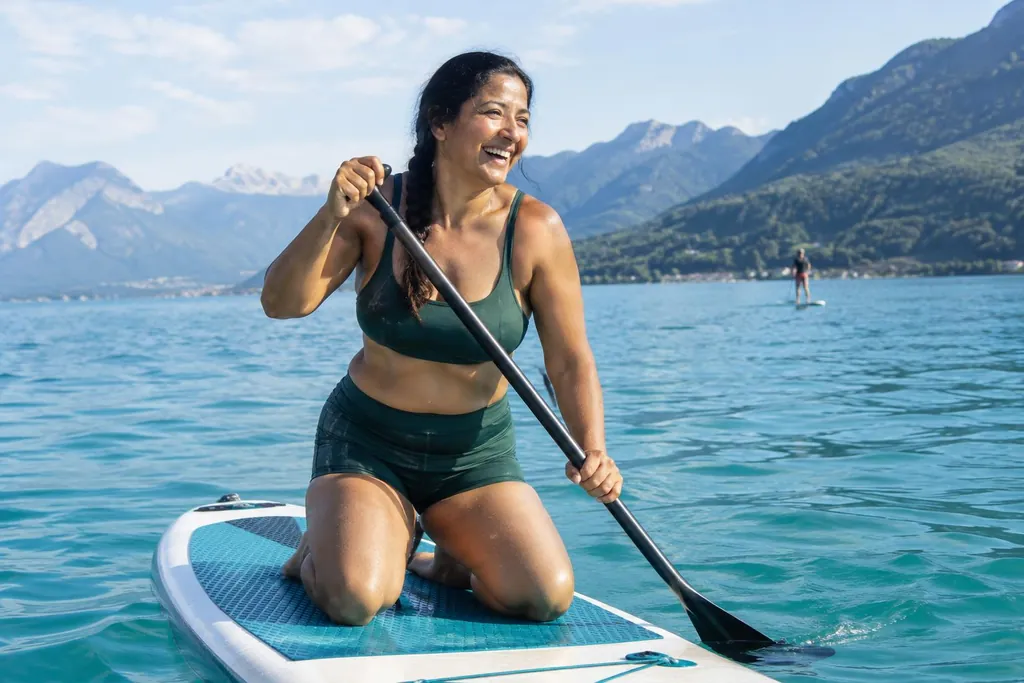 Femme d'environ quarante-cinq ans en brassière de bain et short de bain verts, à genoux sur un paddle sur le lac d'Annecy